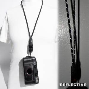 以下が含まれることがあります： 白いドット柄の反射材があしらわれた黒いカメラストラップ。ストラップは黒いキヤノンのカメラに取り付けられており、頑丈な黒いプラスチック製のコネクターが付いています。右下に「REFLECTIVE」という文字が白で印刷されています。