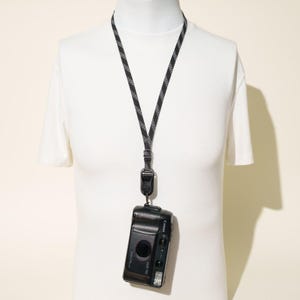 May include: A black Canon Multi Tele film camera hangs from a black and white patterned rope neck strap. The camera features a circular lens, a rectangular viewfinder, and a small flash unit on the front. The strap includes an adjustable buckle.