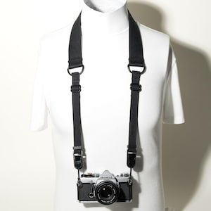 May include: A black camera strap with adjustable metal buckles and D-rings attached to a silver and black Olympus camera. The strap features a wide neck section and narrow webbing that connects to the camera body via secure mounting clips.