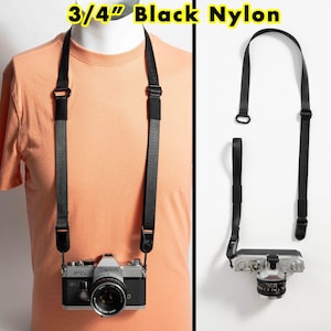 May include: A 3/4 inch black nylon camera strap featuring adjustable sliders and durable attachment clips. The strap is shown attached to a silver and black Canon FTb camera, demonstrating its functional use for carrying equipment.