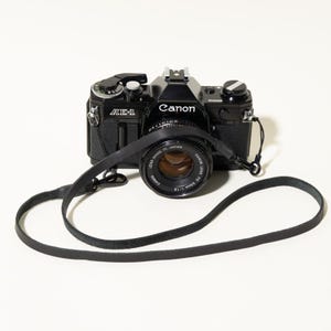 May include: A black Canon AE-1 35mm film camera with a 50mm f/1.8 lens attached. The camera features a silver shutter speed dial, a film advance lever, and a black leather neck strap with metal clips attached to the sides of the camera body.