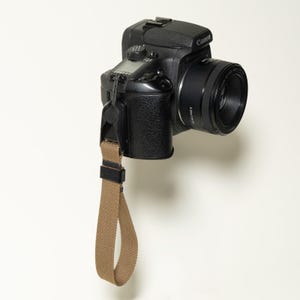 May include: A black Canon digital camera with a lens attached, featuring a tan fabric wrist strap connected to the side. The camera body has a textured grip, a top display screen, and a black plastic connector securing the strap to the device.
