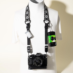 May include: A black Canon AE-1 camera hangs from a black and white patterned woven strap. A small clear film canister and a green and black disposable camera are attached to the strap via black clips, resting against a white shirt.