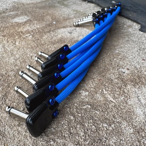 May include: A set of six blue and black instrument patch cables with silver connectors. The cables are braided and arranged in a fan shape, with the connectors neatly aligned. The background is a textured, light-colored surface.