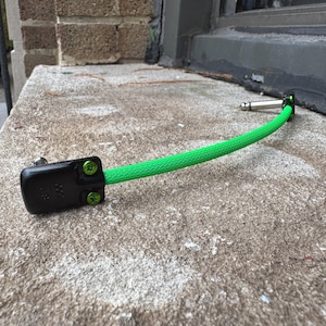 May include: A black guitar pedal connector with green screws is connected to a bright green cable. The cable leads to a silver-colored connector. The items are on a textured, light-colored surface.