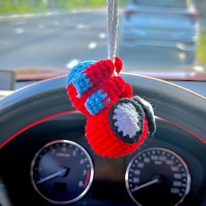 May include: A handmade, red, blue, and white crocheted car charm hanging from a white string. The charm has a round shape with a white and black design on the side. The charm is hanging in front of a car dashboard.