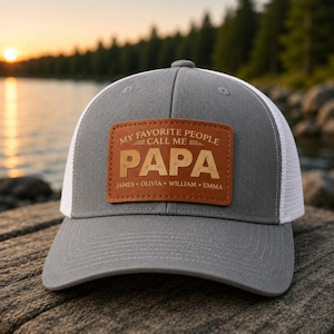 May include: Gray and white trucker hat with a brown leather patch. The patch reads "MY FAVORITE PEOPLE CALL ME PAPA" with the names James, Olivia, William, and Emma below. The hat is on a wooden surface.