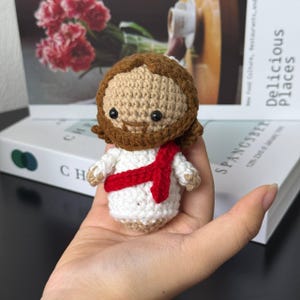 May include: A handmade crochet doll depicting Jesus, featuring a tan face and hair, a brown beard, and a white robe with a red sash. The doll is held in a hand, with a blurred background of books and flowers.