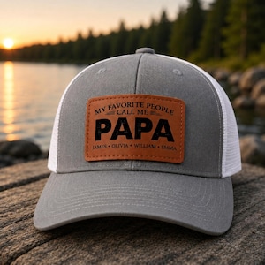 May include: Gray and white trucker hat with a brown leather patch. The patch reads "MY FAVORITE PEOPLE CALL ME PAPA" with the names James, Olivia, William, and Emma below. The hat is on a wooden surface.