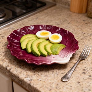 May include: A burgundy-colored, leaf-shaped plate holds sliced avocado and two halves of a hard-boiled egg. A silver fork rests on the countertop next to the plate. The food is arranged on a speckled, light brown countertop.