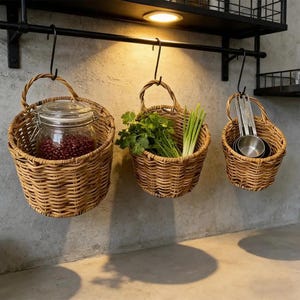 May include: Three hanging woven baskets with handles. One basket holds a glass jar filled with red beans. Another contains green herbs and scallions. The third basket has measuring cups. All are suspended from a black metal bar.