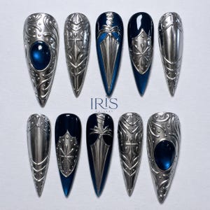 Pode incluir: Um conjunto de dez designs de unhas longas e pontiagudas em prata. Alguns apresentam uma base azul escura com detalhes prateados, outros são inteiramente prateados com uma gema azul. O texto "IRIS NAILARY" é visível. Tema medieval.