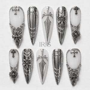 May include: A collection of ten silver-coloured artificial nails, each with a long, pointed shape. The nails display detailed designs such as crosses and fleur-de-lis. They have a white base and are arranged on a white surface.