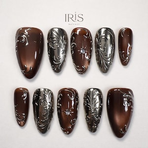 May include: A set of ten artificial nails in a variety of shapes. The nails feature a metallic silver and brown color scheme with ornate, swirling designs. Some nails have a central jewel. The brand name "IRIS NAILXY" is at the top.