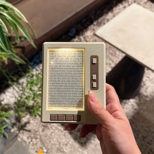 May include: A beige handheld electronic reading device with a rectangular screen displaying Vietnamese text. The device features three vertical buttons on the right side and four horizontal buttons along the bottom edge, all in a matching beige color.