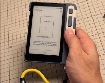 Può includere: Un dispositivo elettronico portatile con una custodia protettiva blu e bianca, dotato di uno schermo che mostra la modalità USB e il testo XTEINK. Un cavo giallo brillante è collegato alla porta inferiore e lo schermo mostra istruzioni di espulsione.