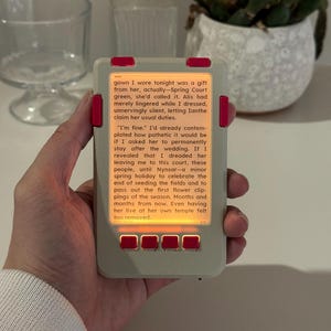 May include: A handheld electronic reading device with a light grey rectangular frame and bright pink accents on the side buttons and bottom controls. The screen displays black text on a warm, orange-tinted background, showing a narrative excerpt.