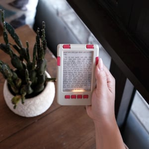 Könnte beinhalten: Ein rechteckiges, handgehaltenes, helles elektronisches Lesegerät mit vier rosa Tasten an der Ober- und Unterkante. Der Bildschirm zeigt einen Block mit vietnamesischem Text. Das Gerät wird aufrecht gehalten und beleuchtet den Text von innen.