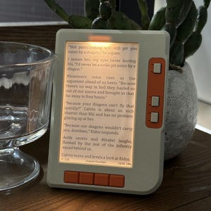Pode incluir: Um leitor de e-book cinza-claro com botões laranja no lado direito e na parte inferior. A tela está iluminada e exibe o texto do capítulo catorze, incluindo diálogos sobre dragões, escribas e uma infantaria, com 32% de progresso.