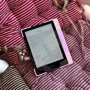 May include: A pink Kindle e-reader with a black screen displaying text, resting on a burgundy and white striped cushion. The e-reader has a light pink border and a lavender accent. The background includes a burgundy and white patterned cushion.