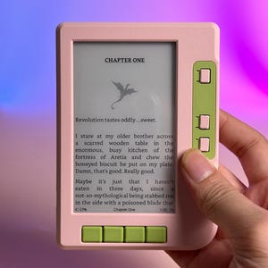 以下が含まれることがあります： 下部に4つ、右側に3つの緑色のボタンが付いたライトピンクの電子書籍リーダー。画面には「CHAPTER ONE」で始まるテキストと小さなドラゴンのアイコンが表示されており、その後に要塞とビスケットに関する段落が続いています。