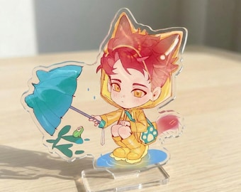 May include: A clear acrylic standee featuring a cartoon character with red hair and fox ears, wearing a yellow raincoat and boots. The character holds a teal umbrella over a small green frog. The design includes a blue puddle base and a paw print bag.