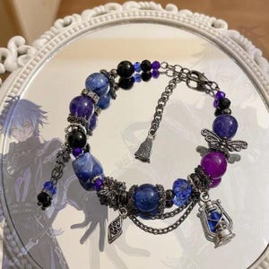 May include: A beaded bracelet featuring blue, purple, and black spherical beads interspersed with silver-toned metal spacers, a small lantern charm, a heart-shaped charm, and a delicate chain link extension with a lobster claw clasp closure.