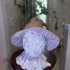 May include: A whimsical, handmade hat with a pointed, tan crown and a wide brim. The brim's underside features a floral pattern with pink flowers on a light blue background. The hat is designed with a ruffled, floral-printed fabric around the base.