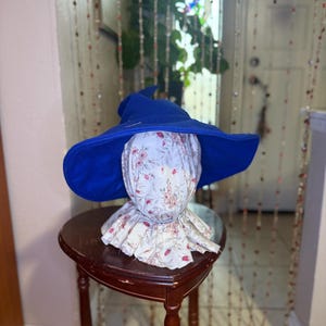 Puede incluir: Un sombrero de bruja azul de fieltro con ala ancha se asienta sobre una cabeza y cuello de tela floral. La cabeza y el cuello están sostenidos por una pequeña mesa de madera oscura. El fondo incluye una puerta y una cortina de cuentas.