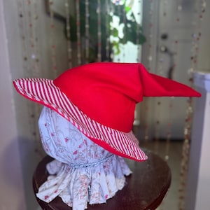 May include: A red hat with a pointed crown and a white and red striped brim. The hat sits atop a floral patterned fabric head form, with a ruffled collar and a blue cord detail. The hat is displayed on a small, dark wooden table.