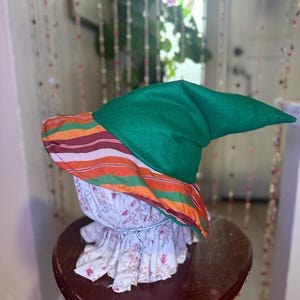 May include: A whimsical decorative hat with a green pointed top and a striped brim in orange, red, and brown. The hat sits atop a floral fabric base, creating a unique and artistic design. The hat is displayed on a small, dark wooden table.