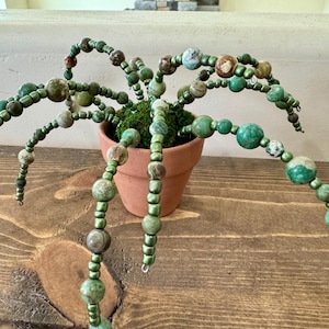 May include: A decorative beaded plant sculpture with green and brown beads, resembling a plant with multiple stems. The sculpture is planted in a small terracotta pot filled with green moss. The beads vary in size and color.