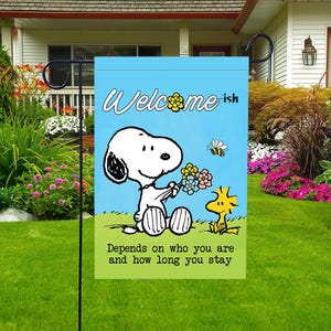 Może przedstawiać: Flaga ogrodowa z Snoopy trzymającym kwiaty, Woodstockiem i pszczołą na jasnoniebieskim tle. Flaga głosi "Welcome-ish" i "Depends on who you are and how long you stay."