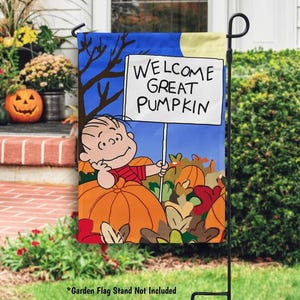 Może przedstawiać: Flaga ogrodowa z kreskówkową ilustracją postaci trzymającej znak z napisem "WELCOME GREAT PUMPKIN". Flaga ma niebieskie tło z dyniami i jesiennymi liśćmi. Czarny metalowy stojak trzyma flagę.