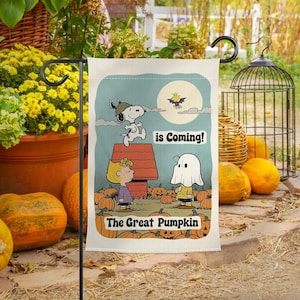 Może przedstawiać: Prostokątna flaga ogrodowa z kreskówkową sceną z Snoopy, Charliem Brownem i postacią ducha. Flaga ma jasnoniebieskie tło z pełnią księżyca i nietoperzem. Tekst brzmi "is Coming!" i "The Great Pumpkin."