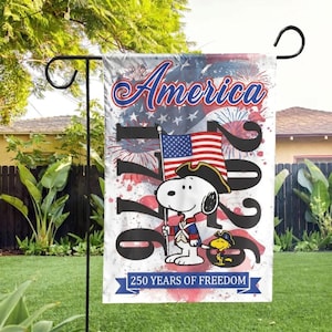 Może przedstawiać: Flaga ogrodowa z Snoopy w mundurze z czasów wojny o niepodległość Stanów Zjednoczonych, trzymający amerykańską flagę. Flaga ma napis "America" w kolorze czerwonym i niebieskim, z napisami "1776-2026" i "250 YEARS OF FREEDOM". Tło ma czerwono-biało-niebieski wzór fajerwerków.