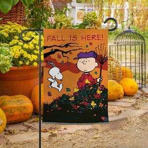 Może przedstawiać: Pomarańczowa flaga ogrodowa z napisem "FALL IS HERE!" z Charliem Brownem i Snoopy'm grabiącymi liście. Flaga ma czarną obwódkę i jest ozdobiona jesiennymi liśćmi i dyniami.