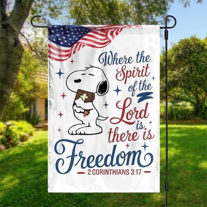 Może przedstawiać: Biała flaga ogrodowa z Snoopy trzymającym książkę, z amerykańską flagą w tle. Flaga zawiera tekst "Where the Spirit of the Lord is, there is Freedom" i "2 Corinthians 3:17."