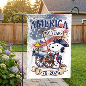 Może przedstawiać: Flaga ogrodowa z Snoopy'm w mundurze z czasów rewolucji amerykańskiej, trzymającym flagę i obok armaty. Flaga zawiera napisy "AMERICA 250 YEARS" i "1776-2026" na tle akwareli z flagą amerykańską.
