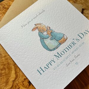 Pu&ograve; includere: Biglietto per la festa della mamma con un'illustrazione ad acquerello di due conigli che si abbracciano. Il biglietto recita "Happy Mother's Day" e "I love you so much already". Il biglietto &egrave; azzurro e bianco con una busta marrone.