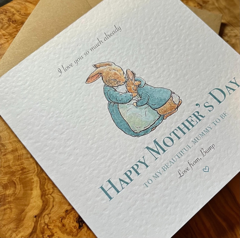 Pu&ograve; includere: Biglietto per la festa della mamma con un'illustrazione ad acquerello di due conigli che si abbracciano. Il biglietto recita "Happy Mother's Day" e "I love you so much already". Il biglietto &egrave; azzurro e bianco con una busta marrone.