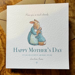 Pu&ograve; includere: Biglietto d'auguri quadrato con un'illustrazione di conigli abbracciati, vestiti di blu. Il testo dice "Buona festa della mamma" e "Ti amo gi&agrave; cos&igrave; tanto". Il biglietto &egrave; su una busta marrone.