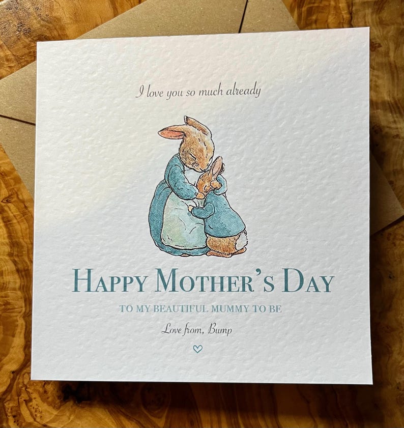 Pu&ograve; includere: Biglietto d'auguri quadrato con un'illustrazione di conigli abbracciati, vestiti di blu. Il testo dice "Buona festa della mamma" e "Ti amo gi&agrave; cos&igrave; tanto". Il biglietto &egrave; su una busta marrone.