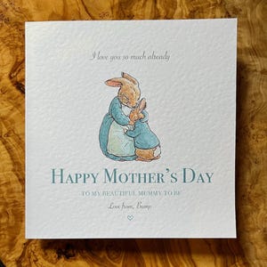 Pu&ograve; includere: Biglietto d'auguri quadrato con un'illustrazione ad acquerello di due conigli che si abbracciano. Il biglietto recita "Happy Mother's Day" e "I love you so much already". Il biglietto &egrave; azzurro e bianco.