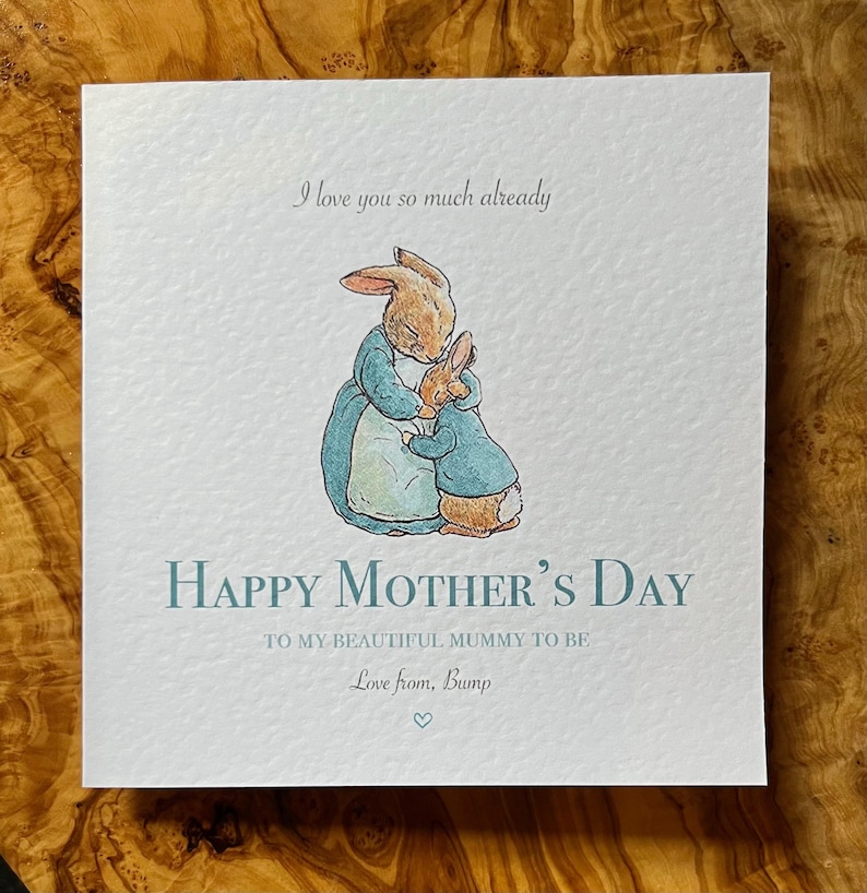 Pu&ograve; includere: Biglietto d'auguri quadrato con un'illustrazione ad acquerello di due conigli che si abbracciano. Il biglietto recita "Happy Mother's Day" e "I love you so much already". Il biglietto &egrave; azzurro e bianco.