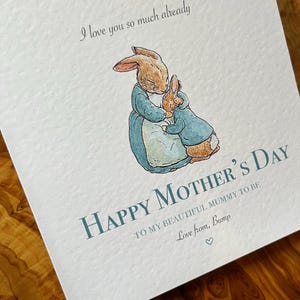 Pu&ograve; includere: Biglietto per la festa della mamma con un'illustrazione ad acquerello di due conigli che si abbracciano. Il biglietto recita "Happy Mother's Day" e include il testo "Ti amo gi&agrave; cos&igrave; tanto" e "Love from, Bump."
