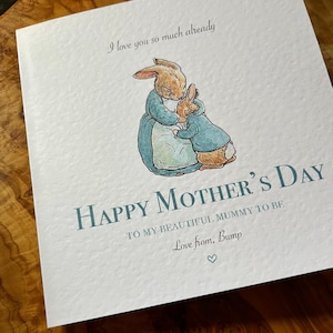 Pu&ograve; includere: Biglietto per la festa della mamma con un'illustrazione ad acquerello di due conigli che si abbracciano. Il biglietto recita "Happy Mother's Day" e "I love you so much already". Il testo &egrave; di colore verde acqua, con un simbolo a forma di cuore in basso.