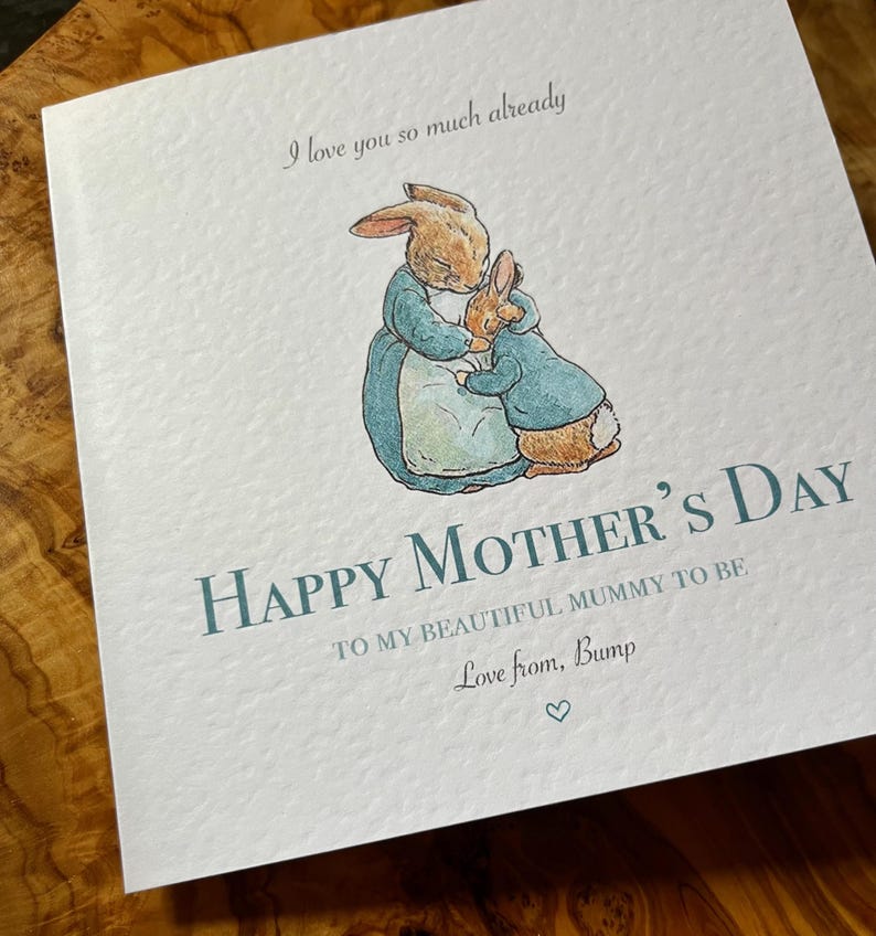 Pu&ograve; includere: Biglietto per la festa della mamma con un'illustrazione ad acquerello di due conigli che si abbracciano. Il biglietto recita "Happy Mother's Day" e "I love you so much already". Il testo &egrave; di colore verde acqua, con un simbolo a forma di cuore in basso.