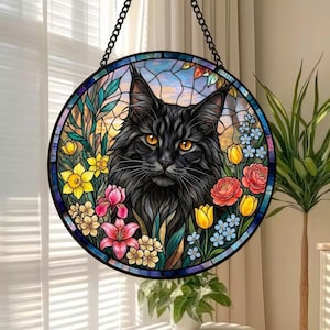 May include: Round stained glass window art featuring a black cat with orange eyes, surrounded by colorful flowers. The cat is the focal point, set against a backdrop of a sunset sky. The artwork is framed by a black metal chain.