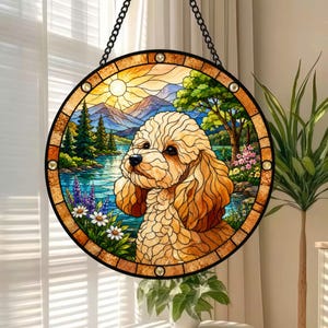 May include: A circular stained glass suncatcher featuring a golden-brown curly-haired dog in the foreground. The background depicts a serene landscape with a blue lake, green pine trees, purple and white wildflowers, and distant mountains under a sun.